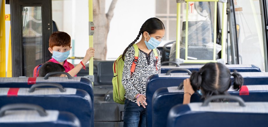 Kids with medical mask coming inside school bus and sitting on seats while maintaining social distance due to coronavirus or covid-19 pandemic - Concept of school reopen or back to school
