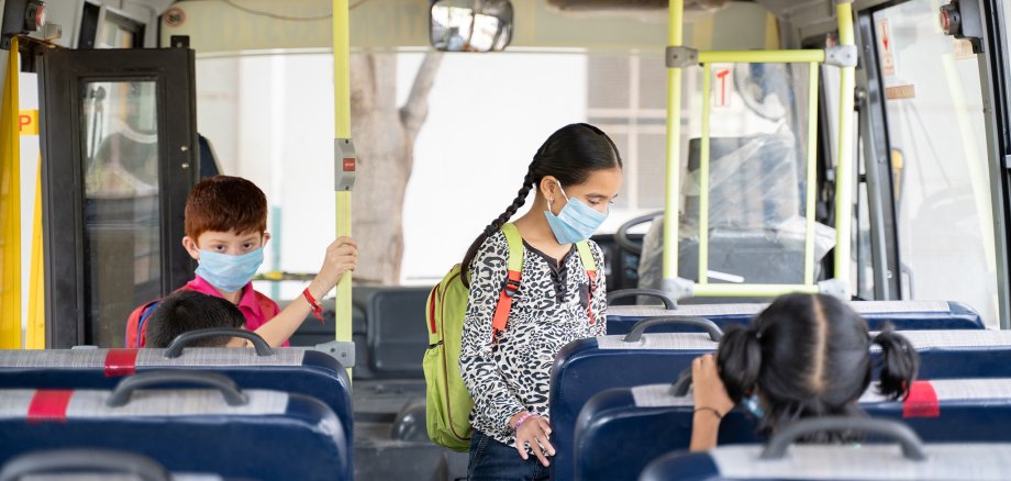 Kids with medical mask coming inside school bus and sitting on seats while maintaining social distance due to coronavirus or covid-19 pandemic - Concept of school reopen or back to school