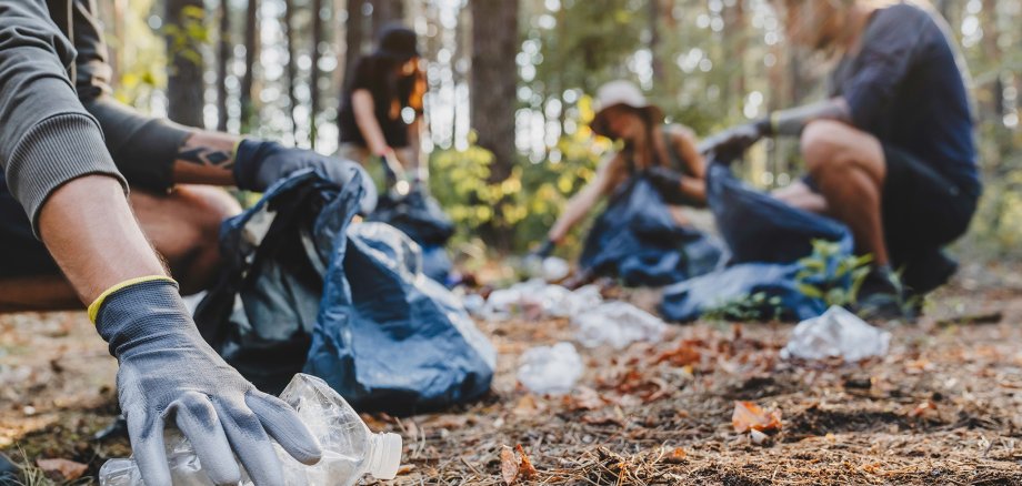 Close up of hands with plastic bottle with group of young people friends volunteers collecting plastic bottles to trash bags in forest background. Ecology concept